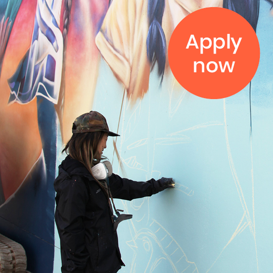 Artist painting on wall with text 'Apply Now'
